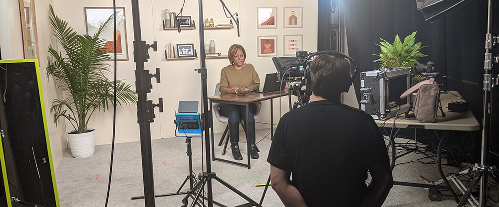 A person sits at a desk in a production studio and talks to a camera. A videographer kneels behind the camera. How to prepare for an interview is one of the top video production FAQs.