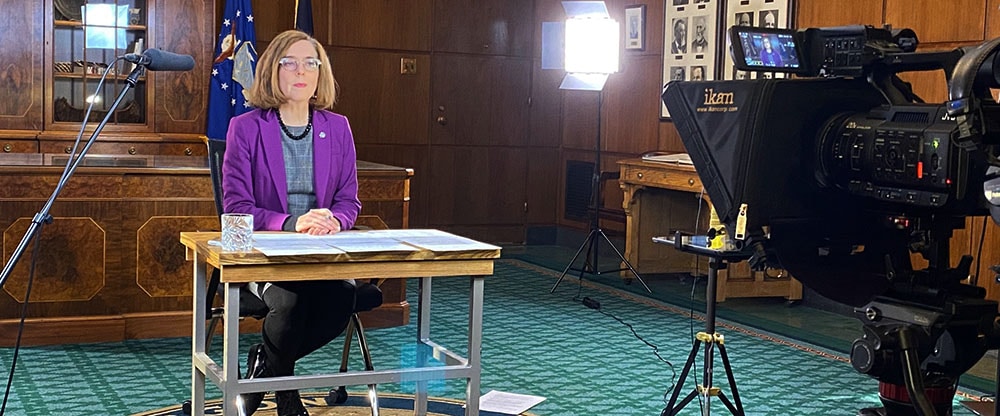 Oregon's Governor Kate Brown is shown sitting behind a table facing a camera during a virtual press conference.
