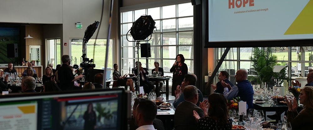 Julie Tolliver Auflick, Chair of the YWCA Board of Directors, hosts the Inspire Hope hybrid event. She is standing in front of a large screen with the event logo. There is a camera in front of her to livestream the event online.