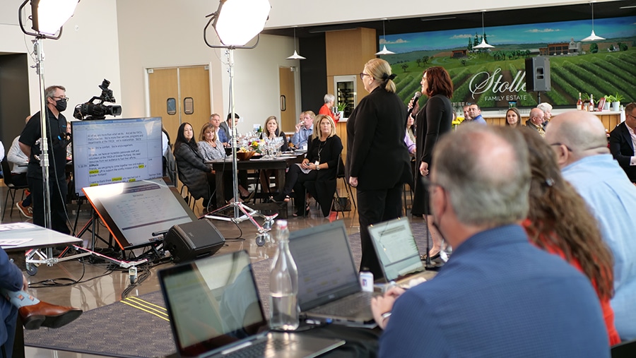 Two people host a hybrid event. Between them and the in-person audience are large lights, a camera and two large monitors. Behind and to the side of the hosts are three people on laptops managing the script and online bidding