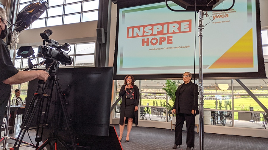 Two event hosts stand in front of a camera, lights, and monitors during a hybrid event.