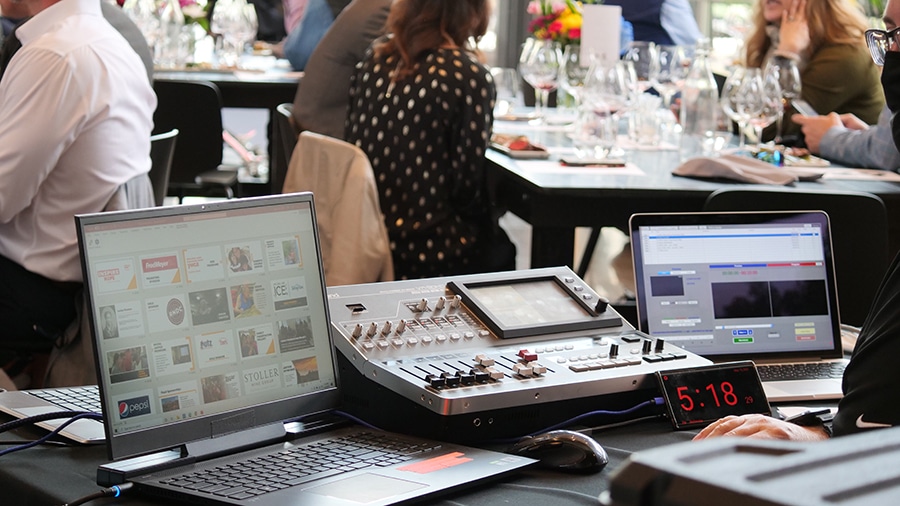 Equipment to run a slide presentation during a hybrid event is set up on a table. Event attendees are at tables in the background