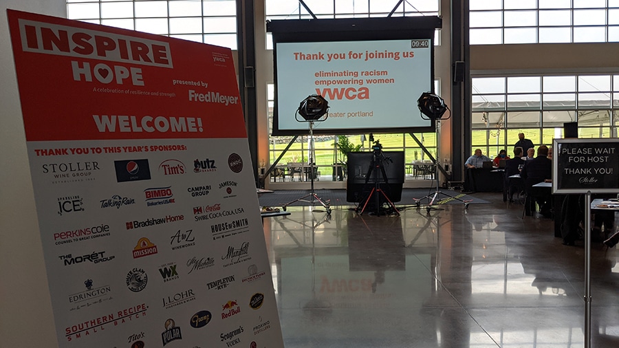 At the entrance to a large event space is placed a welcome sign thanking even sponsors. The event space has a wall of windows ad overlooks a large green lawn.