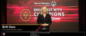 A moment from the Special Olympics Oregon Breakfast with Champions livestream shows the host in front of a large LED wall. Overlayed on the video is the nameplate and captions