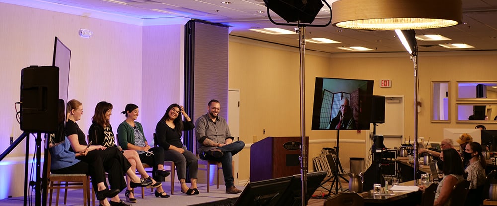 Six panelists speak from the stage when another panelis is seen on a large monitor during a hybid conference