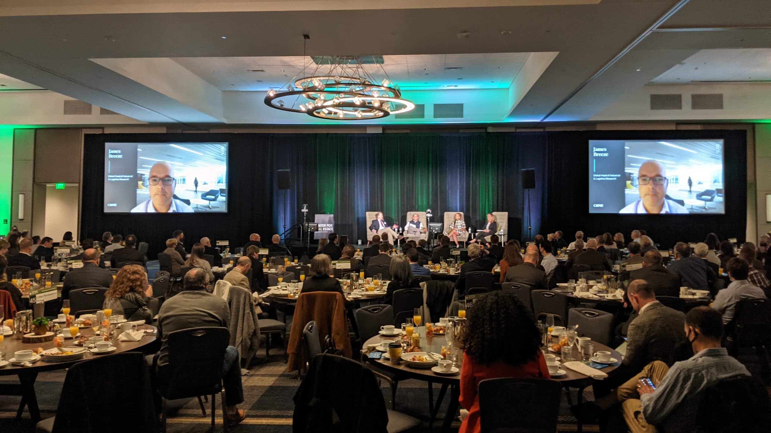 In-Room Audience View of a Hybrid Conference. The live view of the remote presenter is shown on the screens as he interacts with the other panelists on the stage. Portland State University Center for Real Estate Annual Real Estate Conference hosted at the Hyatt at the Oregon Convention Center.