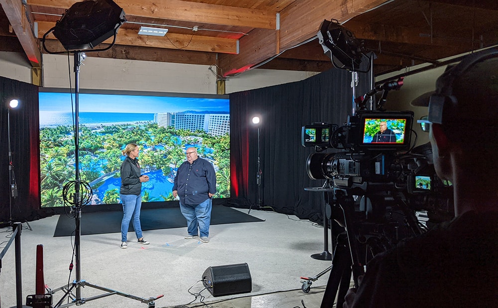 Two hosts stand in front of the camera during a virtual event live stream. A camera operator looks at the framed image through the camera lens