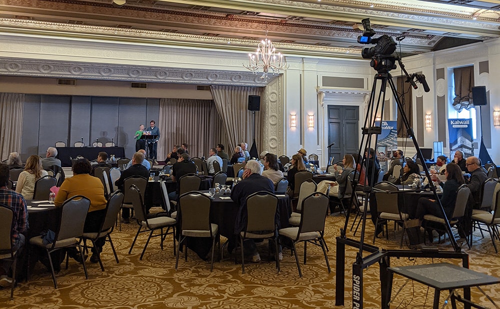 A camera in the back of a hotel ballroom captures the presentations for a live stream of the event