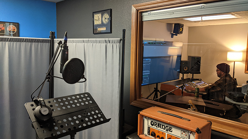 Voice over production studio at The AV Department. A stand, headphones, and microphone are positioned by a window looking into an audio recording suite. The audio engineer is sitting at a mixing console
