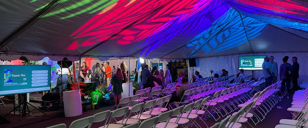 Colorful lighting spans the inside top of a large white event tent. White chairs are set up for guest seating.