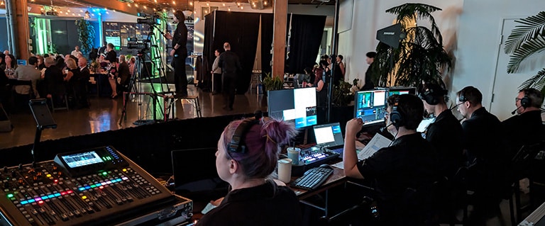 An AV production team operating audio and video equipment at a live event are seated at the tech table. Multiple monitors, a mixing console, other AV gear are visible. A camera operator and part of the audience are visible in the background.