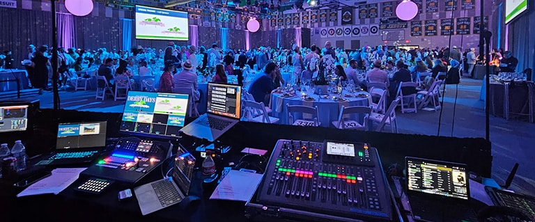 View from the AV tech table. The audio mixing console, computers and monitors are visible during a fundraising gala.