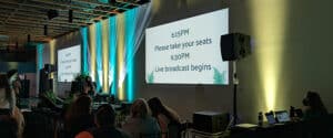 People are seated in an event space with a screen showing: "6:15 PM Please take your seats. 6:30 PM Live broadcast begins." Colorful lights illuminate the wall. Looping slide shows and lighting cues are two actions to include in a Run of Show for you event.